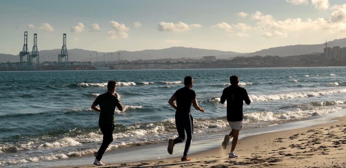 Three men jogging along a scenic beach, embracing a healthy lifestyle under a clear sky.