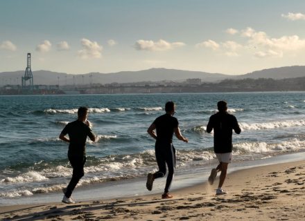 Three men jogging along a scenic beach, embracing a healthy lifestyle under a clear sky.
