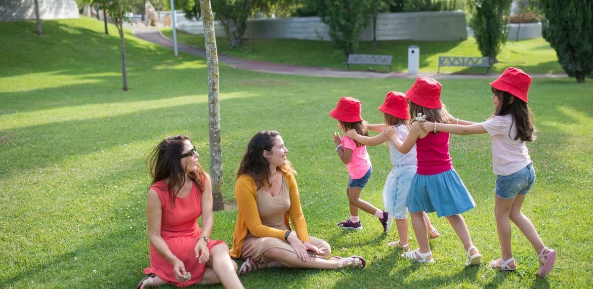 Happy children playing in a sunny park with two mothers sitting nearby, enjoying the outdoors.