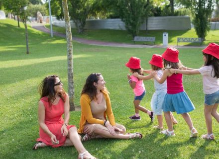 Happy children playing in a sunny park with two mothers sitting nearby, enjoying the outdoors.
