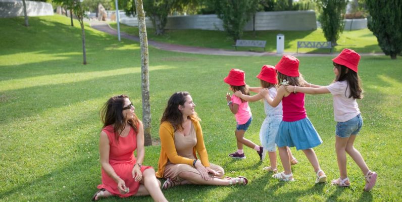 Happy children playing in a sunny park with two mothers sitting nearby, enjoying the outdoors.