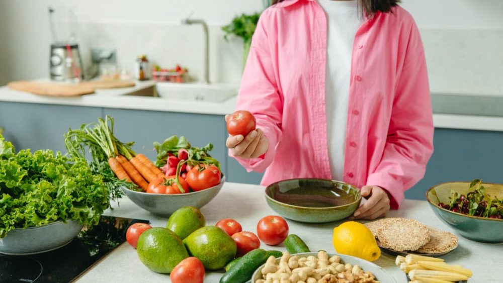 Woman in pink overshirt holds tomato in kitchen with fresh fruits and veggies.