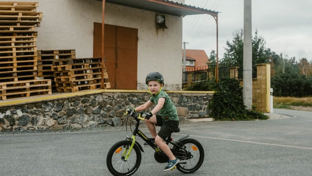 A young boy in a helmet rides his bicycle near a warehouse with stacked wooden pallets.